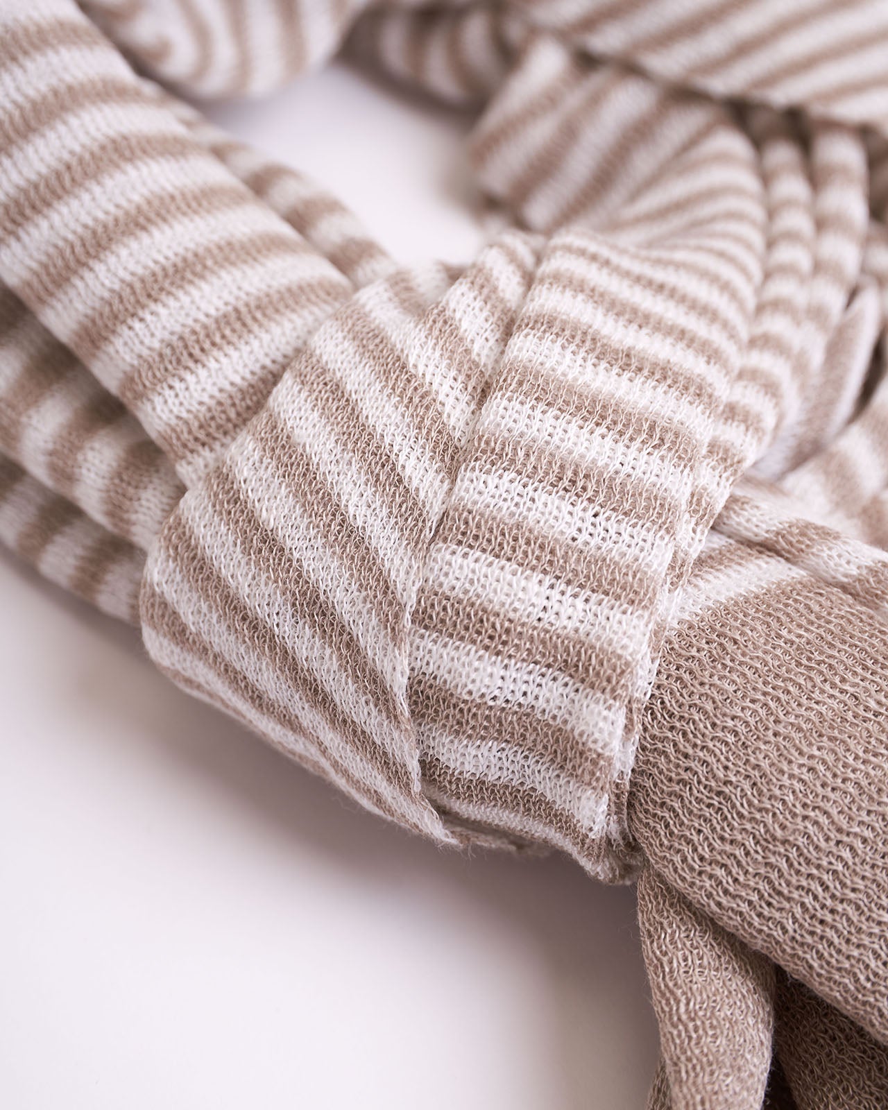 Soft textured linen scarf arranged in a loose knot, featuring a delicate beige and white striped pattern with subtle textural details. The ends of the scarf have a frayed, fuzzy finish, adding a casual yet elegant touch. The scarf is displayed against a white background, highlighting its cozy and inviting appearance