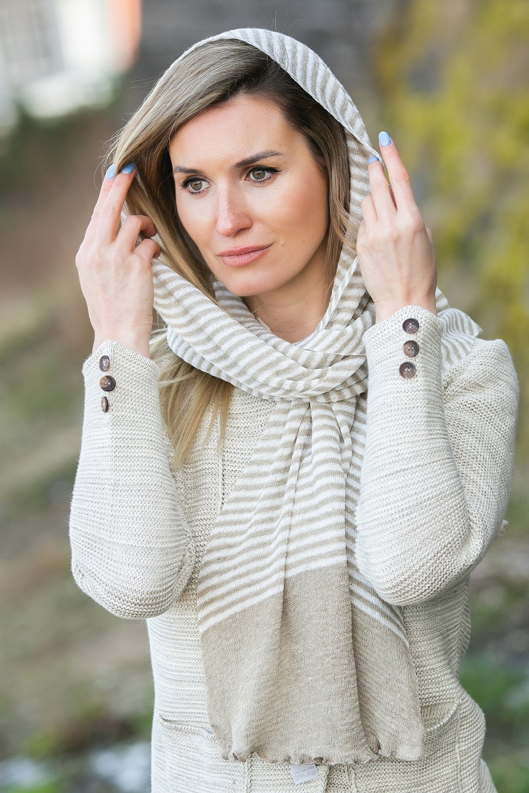 Elegant woman wearing a stylish linen beige striped scarf and textured sweater, ideal for fall fashion. The scarf is loosely wrapped around her neck, extending over her head, accentuating her blonde hair and contemplative expression. Her look is completed with a light beige and white striped pattern, perfect for a chic autumn outfit. The soft, blurred background features green foliage, adding a natural touch to the scene