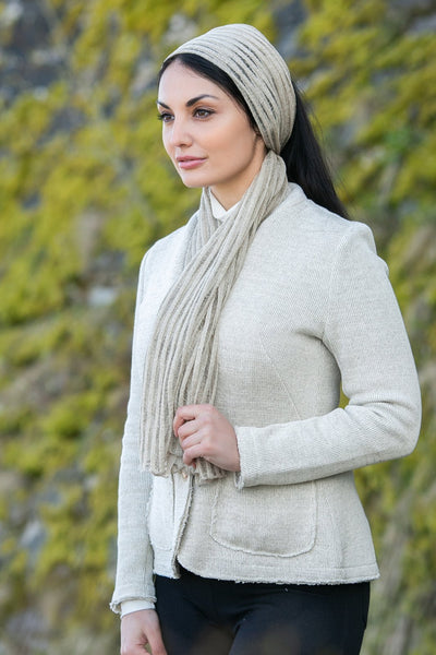Elegant woman outdoors, showcasing a linen scarf wrapped stylishly around her head and neck. The scarf's fine ribbed texture and natural beige color highlight its quality and simplicity. Her serene expression and the soft green foliage background emphasize the scarf's organic and earthy appeal, perfect for a chic, sustainable fashion statement